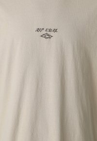 Bež majica od pamuka s okruglim izrezom, s crnim izvezenim natpisom i logom "RIP CURL" na gornjem prednjem dijelu.