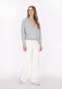 Grauer gestrickter Pullover mit V-Ausschnitt, langen Ärmeln und enganliegendem Taillenbund, kombiniert mit weißen weiten Hosen und Canvas-Sneakern.