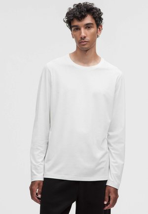 Jonge man met krullend haar die een effen wit shirt met lange mouwen en zwarte broek draagt, staand tegen een lichtgrijze achtergrond.