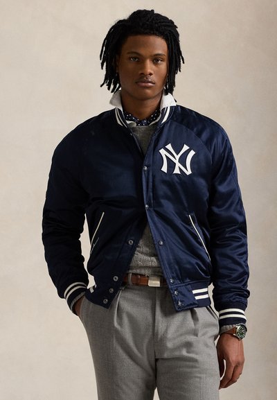 Fiatal férfi dreadlock hajjal, sötétkék New York Yankees dzsekit, szürke pulóvert, szürke nadrágot és karórát visel, semleges háttér előtt áll.