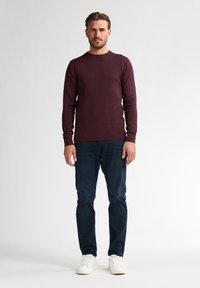 Bordeauxroter Rundhals-Pullover aus weichem Stoff, kombiniert mit dunkelblauen Jeans und weißen Sneakers, vor einem einfarbigen Hintergrund stehend.