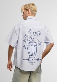 Camicetta a maniche corte a righe in azzurro chiaro e bianco, con una grafica di un vaso floreale e testo sul retro, realizzata in tessuto di cotone.