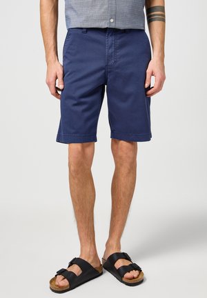 Navyblaue Shorts aus Baumwolle mit geradem Schnitt, seitlichen Taschen und umgeschlagenen Säumen, kombiniert mit schwarzen Sandalen mit verstellbaren Riemen.