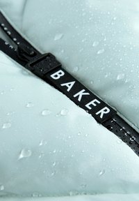 Vodoodporna svetlo modra tkanina z vodnimi kapljicami, opremljena s črnim trakom, označenim "BAKER", in plastičnim sponko.