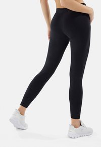Leggings neri elasticizzati con vita alta, texture liscia e gambe affusolate, abbinati a sneakers sportive bianche.