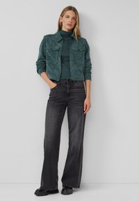 Turkusowa kurtka z kordurowego materiału z dwiema kieszeniami na piersi, zestawiona z ciemnozielonym golfem i czarnymi dżinsami z rozszerzanymi nogawkami. Czarne buty na platformie dopełniają całość.