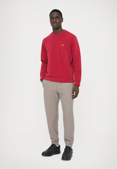 Κόκκινο καπιτονέ φούτερ με γύρο λαιμόκοψη, μικρό λογότυπο στο στήθος· συνδυασμένο με ανοιχτό γκρι υφασμάτινο παντελόνι και μαύρα αθλητικά παπούτσια.