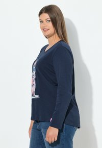 Marineblaue Langarm-T-Shirt mit V-Ausschnitt, versehen mit einem bunten Hasenmotiv auf der Vorderseite und einem abgerundeten Saum. Kombiniert mit blauen Jeans.