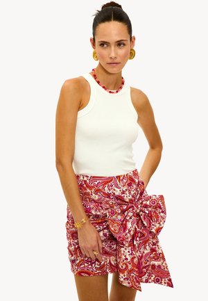 Un débardeur blanc à côtes associé à une jupe enveloppante à motif paisley rose ornée d'un grand nœud à la taille. Des accessoires dorés complètent la tenue.