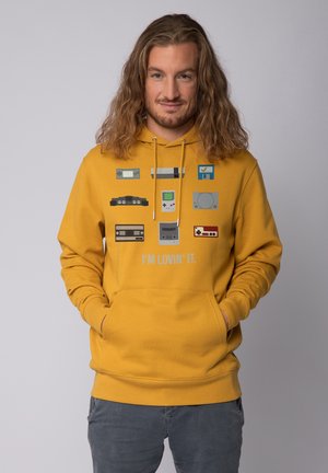 Sweat-shirt à capuche jaune avec des consoles de jeux rétro et des manettes imprimées, accompagné d'un texte gris qui dit "JE L'ADORE." Poche kangourou à l'avant.
