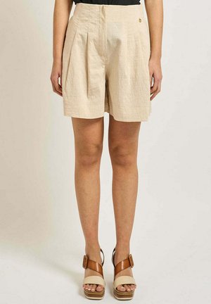 Short beige plissé taille haute porté avec des sandales compensées marron clair et beige sur une personne debout devant un fond uni.