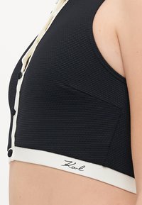Black cropped top with a textured fabric, featuring a white button placket and logo accent at the hem. The design includes side pockets.