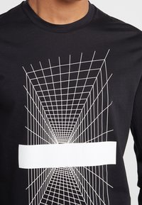 Svart långärmad t-shirt med ett vitt geometriskt rutnät och ett solid horisontellt vitt panel på bröstet.