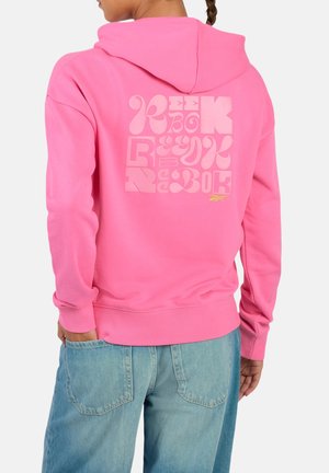 Persoon met een roze hoodie met gestileerde "Reebok" tekst op de achterkant en een blauwe spijkerbroek, van achteren weergegeven tegen een effen achtergrond.