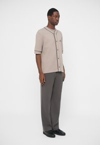 Camicia beige a maniche corte con bottoni e bordo nero; abbinata a pantaloni larghi grigi scuri e sneakers nere. Design semplice e casual.