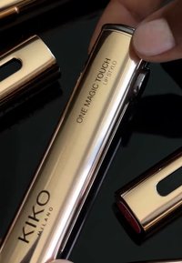 Guldmetallisk läppstiftbehållare märkt "ONE MAGIC TOUCH LIP STYLO," med en glansig yta och en liten öppning som avslöjar en färgad ände, på en svart yta.