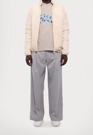 Mies, joka käyttää beigeä toppatakki, harmaat leveälahkeiset housut, beige t-paita sinisellä kuviolla ja valkoiset lenkkarit, seisoo valkoista taustaa vasten.