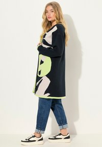 Cardigan lavorato a maglia di colore blu scuro con motivi astratti verdi e beige, dotato di orlo arrotondato e maniche lunghe, abbinato a jeans arrotolati e sneaker.