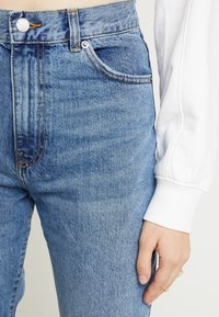 Högmidjade blå denimjeans med ljus tvätt, enkel metallknappstängning och femficksdesign. Har synlig sömnad och mjuk textur.