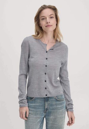 Grijze gebreide cardigan met lange mouwen, ronde hals en zwarte knopen aan de voorkant. Draag over lichtblauwe spijkerbroek. Glad oppervlak.