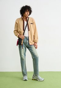 Beige fleece jacket with a maroon zipper and accents, paired with light blue jeans and gray sneakers. Small black crossbody bag featured.