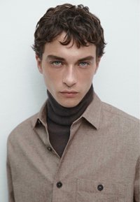 Brun knap-op skjorte med en textured finish, båret over en mørk turtleneck. Modellen har krøllet hår og lyseblå øjne, med en neutral baggrund.