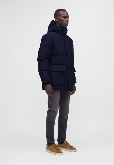 Homme portant une doudoune bleu marine avec capuche, un jean gris et des baskets lacées couleur beige, debout légèrement de profil à droite sur un fond uni.