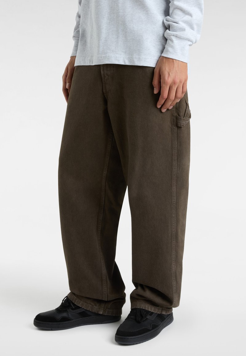 Brown baggy trousers with a smooth texture and side pocket. Paired with black sneakers, the outfit features a light gray long-sleeve top.
