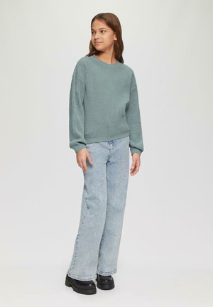 Getexturede, groene gebreide trui met lange mouwen, gecombineerd met lichtblauwe uitlopende spijkerbroek en chunky zwarte platform schoenen.