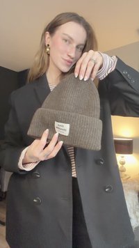 Brown ribbed knit beanie with a folded cuff and a tag labeled "MADS NØRGAARD" displayed by a person wearing a black blazer and striped shirt.