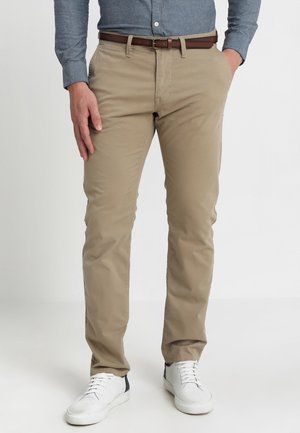 Mand iført beige chinos, brun bælte, blå skjorte med knapper og hvide sneakers, stående med den ene hånd i lommen mod en hvid baggrund.
