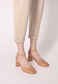 Béžové lakované kožené sandály na kotníku se čtvercovou špičkou a blokovým podpatkem. Hladký povrch s jemným leskem a nastavitelné detaily pásku.