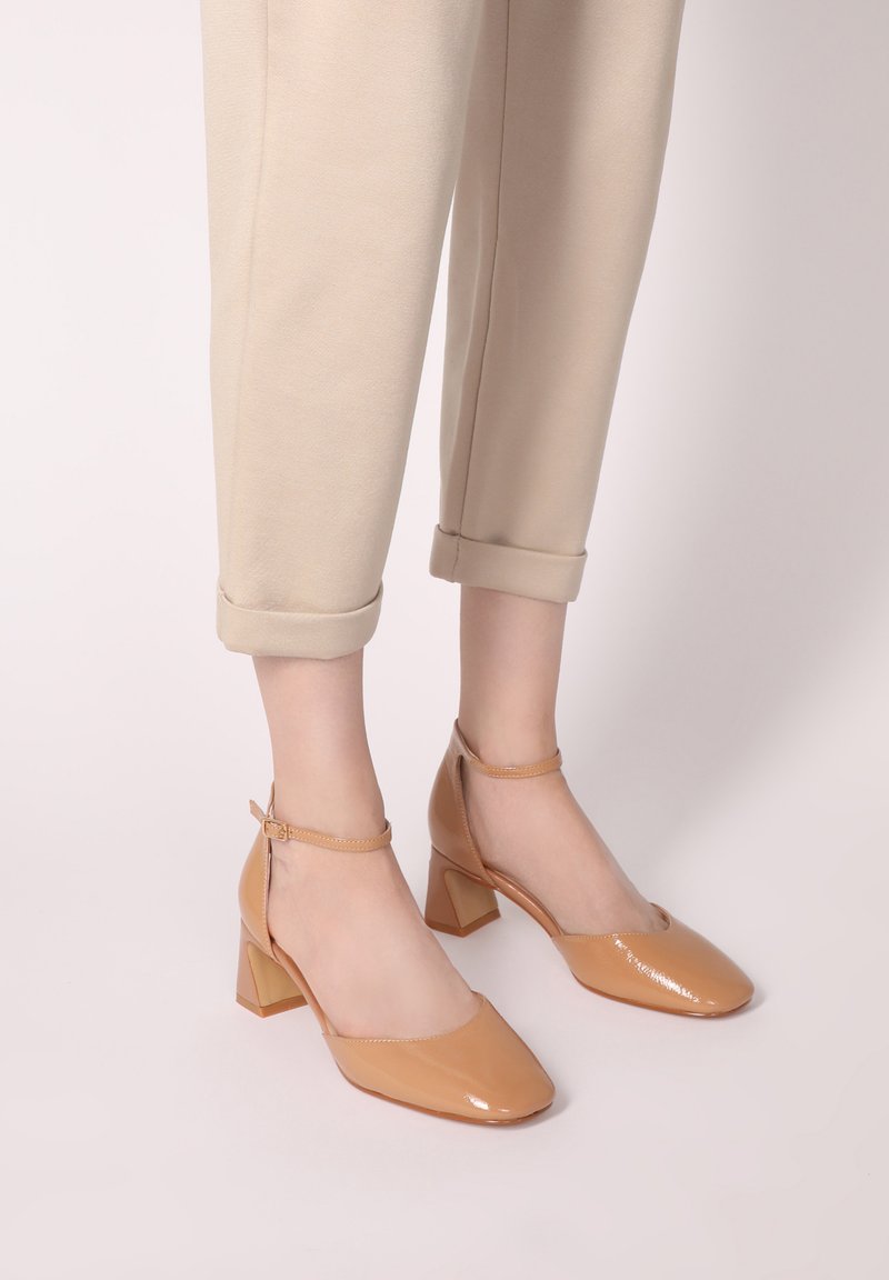 Béžové lakované kožené sandály na kotníku se čtvercovou špičkou a blokovým podpatkem. Hladký povrch s jemným leskem a nastavitelné detaily pásku.