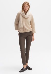 Beige ribbed sweater with wide sleeves, paired with dark brown fitted pants and black flat shoes. The scarf features soft fringe detailing.