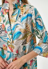 Seidenshirt mit floralem Muster in Creme, versehen mit lebhaften Blau-, Grün- und Rottönen, einem Stehkragen und drei Viertel langen Ärmeln.