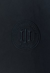 Černá tkanina s texturovaným povrchem, na které je vyražené kruhové logo s nápisem "TOMMY HILFIGER 1985" ve středu.