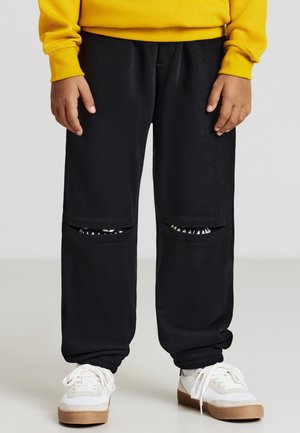 Bambino che indossa pantaloni neri con ritagli sulle ginocchia che mostrano un motivo a denti di squalo sotto, sneakers bianche e una felpa gialla.