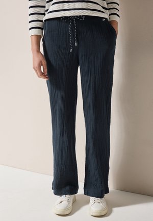 Kobieta ubrana w teksturowane granatowe spodnie z troczkami i kieszeniami, białe sneakersy oraz pasiasty, długorękawowy top, stojąca na tle beżowej ściany.