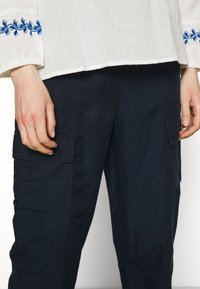 Biała lniana bluzka z niebieskimi haftowanymi akcentami na rękawach, w połączeniu z granatowymi spodniami cargo z dwoma bocznymi kieszeniami i luźnym krojem.