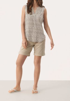 Femme portant un haut beige sans manches à motifs, un short kaki clair retroussé et des sandales blanches façon tongs, debout devant un fond uni.