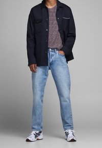 Veste marine avec poches, t-shirt gris texturé, jean bleu clair et baskets blanches avec des accents noirs. Tenue décontractée en plusieurs couches.