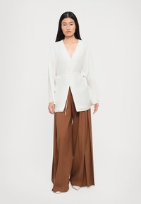 White long-sleeve blouse with deep V-neck and tie waist, paired with wide-leg brown trousers. Smooth fabric with minimal detailing.