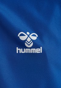 Blåt stof med en glat tekstur og et fremtrædende hvidt Hummel-logo, der inkluderer et abstrakt bi-symbol og stiliseret tekst.