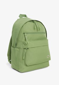 Sac à dos vert avec poches zippées à l'avant et sur le dessus, poche latérale, bretelles rembourrées et petit logo crocodile brodé dans le coin de la poche avant.