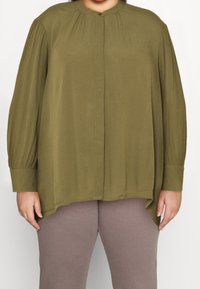 Blouse à manches longues vert olive avec fermeture à boutons, épaules plissées, ourlet rond et texture douce, associée à des leggings marron côtelés.