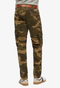 Man wearing green and brown camouflage cargo pants with flap pockets, a brown belt, gray shirt, and white sneakers.