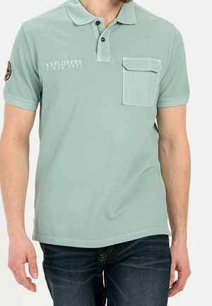 Mężczyzna w jasnozielonej, krótkorękawowej koszulce polo z przednią kieszenią, rozpinanym kołnierzykiem i napisem "EXPLORERS SINCE 1977" na piersi.