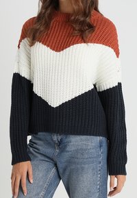 Sweter dziany w kolorach rdzawego, białego i granatowego z wzorem w jodełkę. Prążkowana faktura, długie rękawy i luźny fason. Swobodny styl z widocznymi szwami.