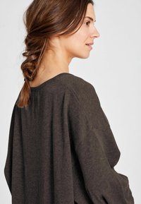 Ženska z rjavimi kitkami, oblečena v ohlapen temno siv pleteni pulover, obrnjena desno proti enostavni svetli ozadju.