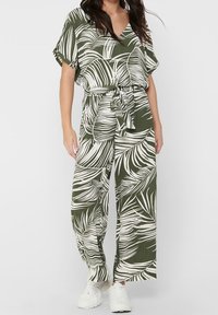 Groene en witte jumpsuit met een bladerenpatroon, met een ontspannen pasvorm, korte mouwen, een strikceintuur en wijde broekspijpen, gedragen met witte sneakers.